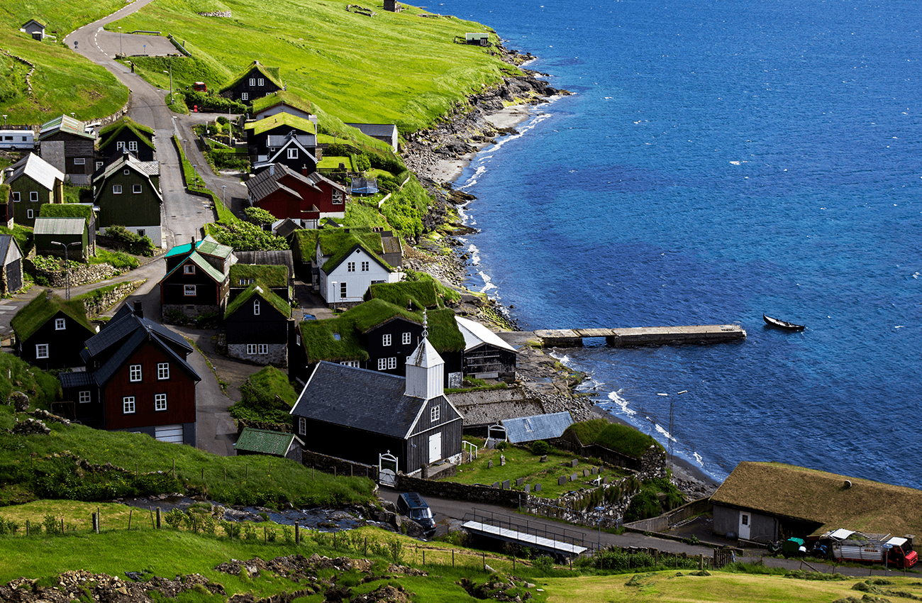 Billede af en kirke og sorte huse ved vandet på grønt og bakket landskab på Færøerne.