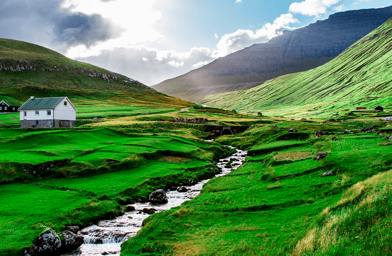Billede af et hus, der ligger ved en å i et grønt bjergrigt landskab på Færøerne.