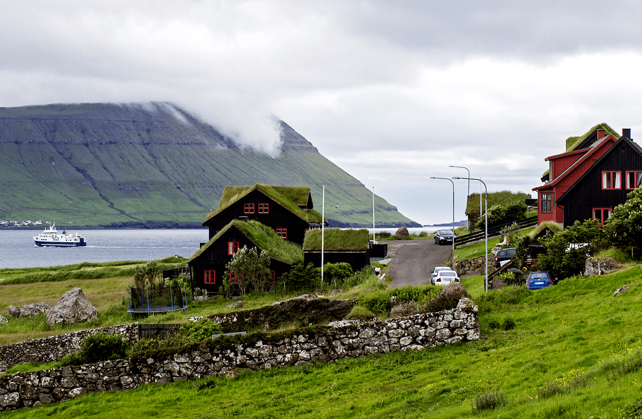 Billede af nogle huse med mos på tagene i Kirkjubøur på Færøerne, med bjerge i baggrunden.