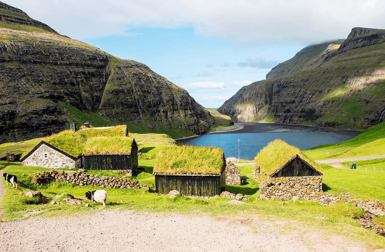 Billede af nogle gamle huse med grønt græs på tagene og med udsigt over en sø med store bjerge rundt om søen på Færøerne.