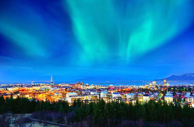 Billede af en oversigt over Reykjavik om aftenen med flotte nordlys over den Islandske hovedstad og bjerge i baggrunden.