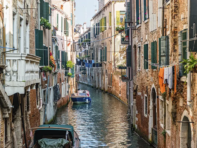 Smal kanal i Venedig med gamle huse, grønne skodder, vasketøj og både i vandet.